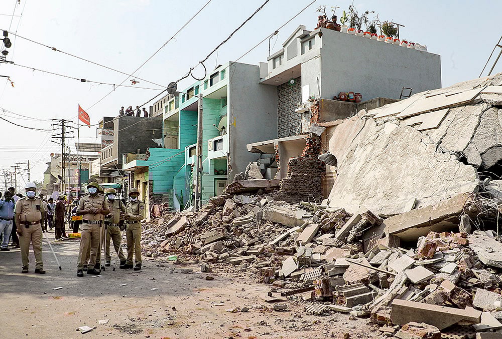 | Photo: PTI : Police personnel stands guard as a demolition drive is underway to clear illegal houses along the Aji river banks and TP Road in the Jangleshwar area, in Rajkot, Gujarat.