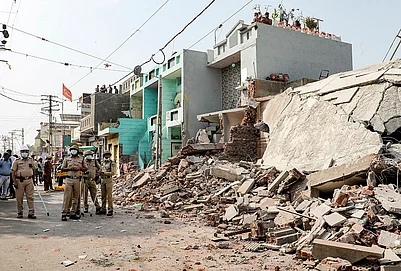 | Photo: PTI : Police personnel stands guard as a demolition drive is underway to clear illegal houses along the Aji river banks and TP Road in the Jangleshwar area, in Rajkot, Gujarat.