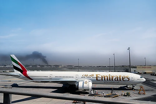 | Photo: AP : Enveloped by Darkness: A plume of smoke caused by an Iranian strike is seen in the background of an Emirates plane parked at the Dubai International Airport on March 1, 2026